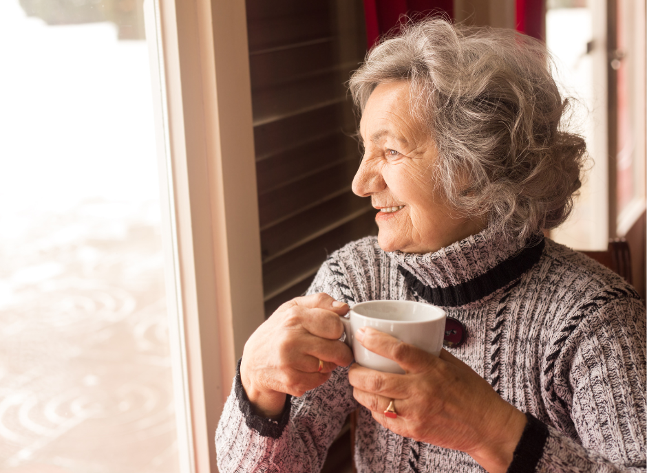 An older woman drinking a cup of coffee in front of a window.