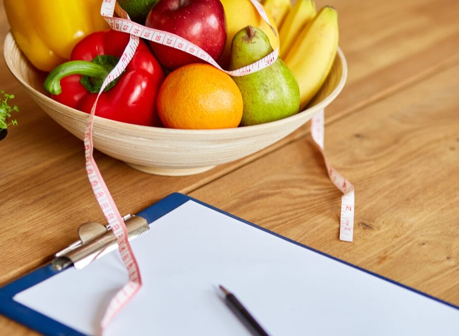 A bowl of fresh fruits and vegetables next to a clipboard with a sheet of paper titled "How to Find a Renal Dietitian" and a measuring tape on a wooden table.