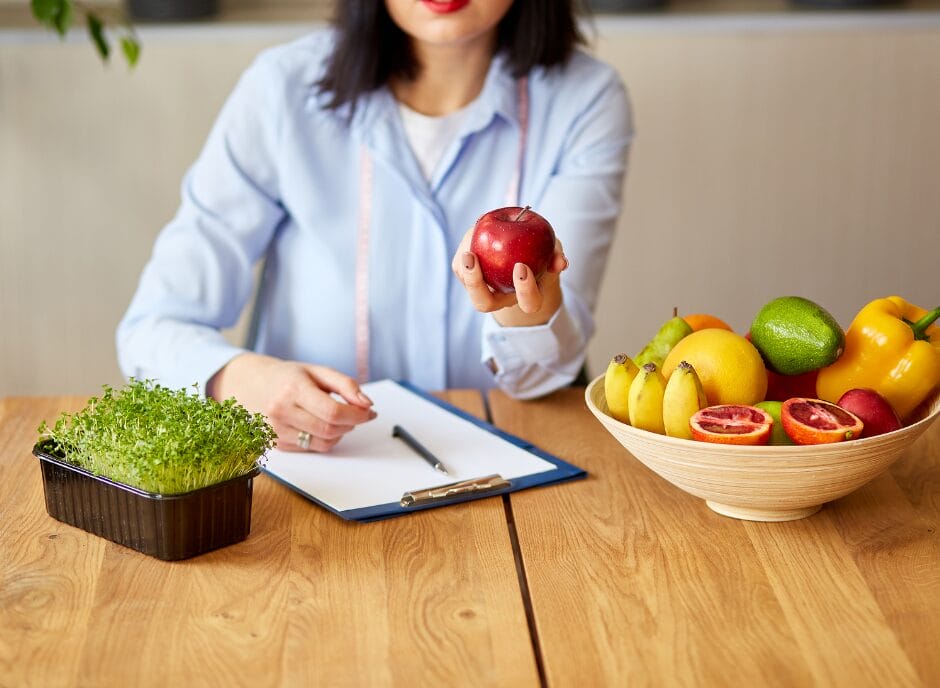 Woman in a blue shirt sitting at a desk, holding a red apple, with a clipboard titled "How to Find a Renal Dietitian" and a bowl of various fruits in front of her.