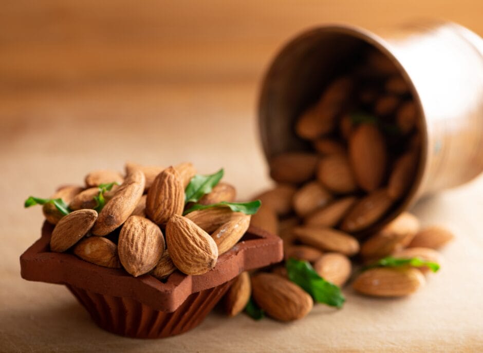 A small wooden bowl filled with almonds is placed on a table, with more almonds spilling out of a tipped-over metal container nearby. Green leaves are scattered among the almonds.