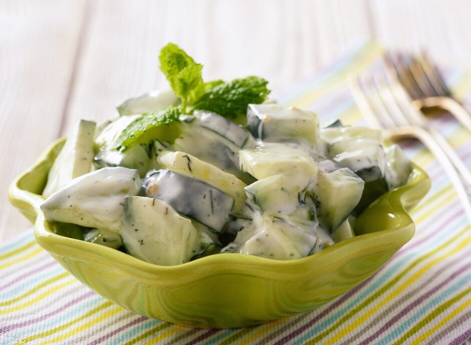 A bowl of cucumber salad with creamy dill dressing, garnished with mint leaves, presented in a green leaf-shaped dish on a striped tablecloth.