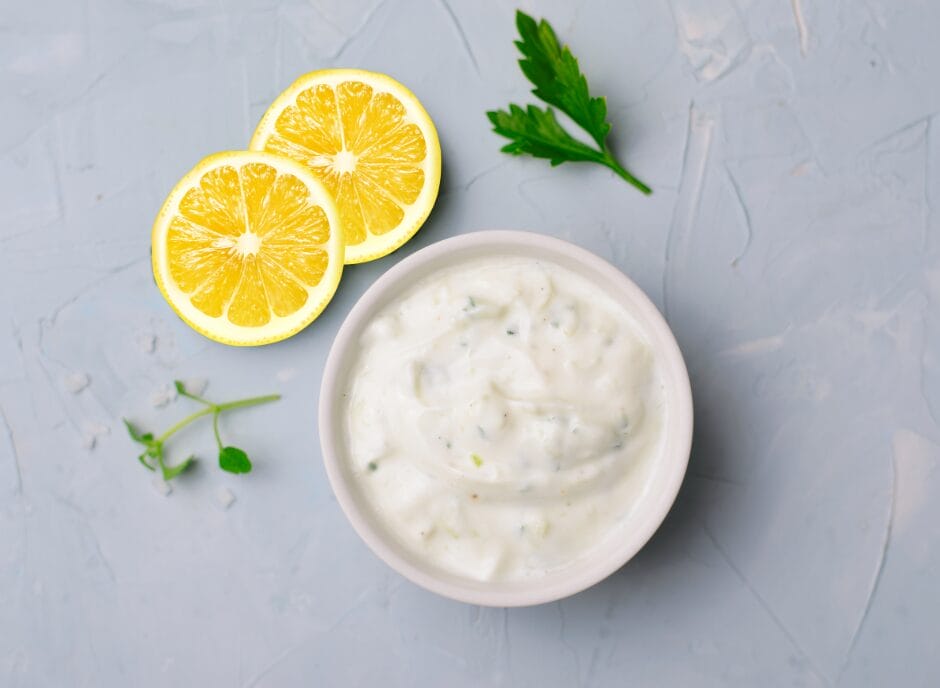 A bowl of creamy white yogurt-based dip with herbs, garnished with two lemon slices and a sprig of parsley on a gray textured background.