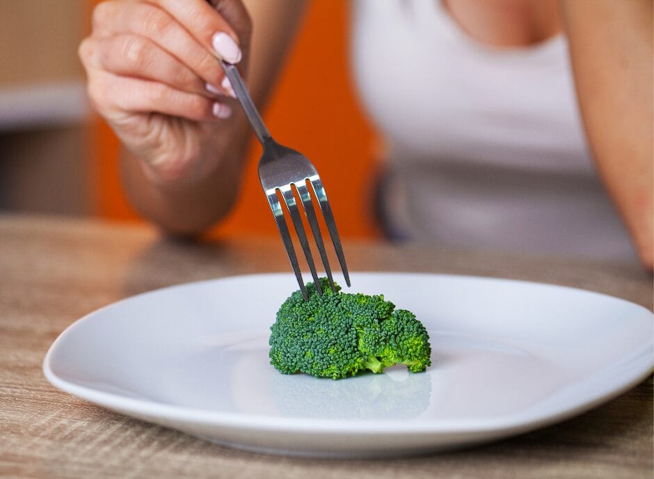 A person wearing a white tank top is holding a fork and spearing a piece of broccoli on a white plate.