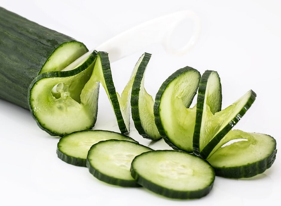 A cucumber partially peeled and sliced on a white surface, with a peeler resting beside it.