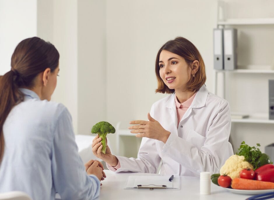 A nutritionist in a white lab coat discusses healthy eating with a woman, using a piece of broccoli as a visual aid, at a desk with various vegetables.