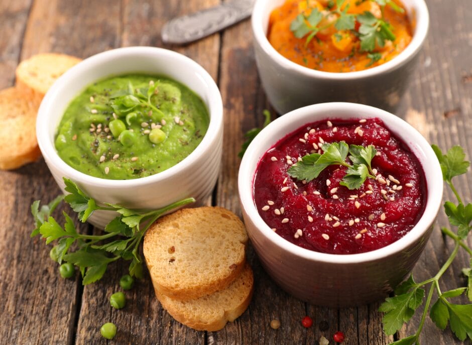 Three bowls of colorful vegetable dips including green, orange, and red, each garnished with herbs, are placed on a wooden table alongside toasted bread slices.