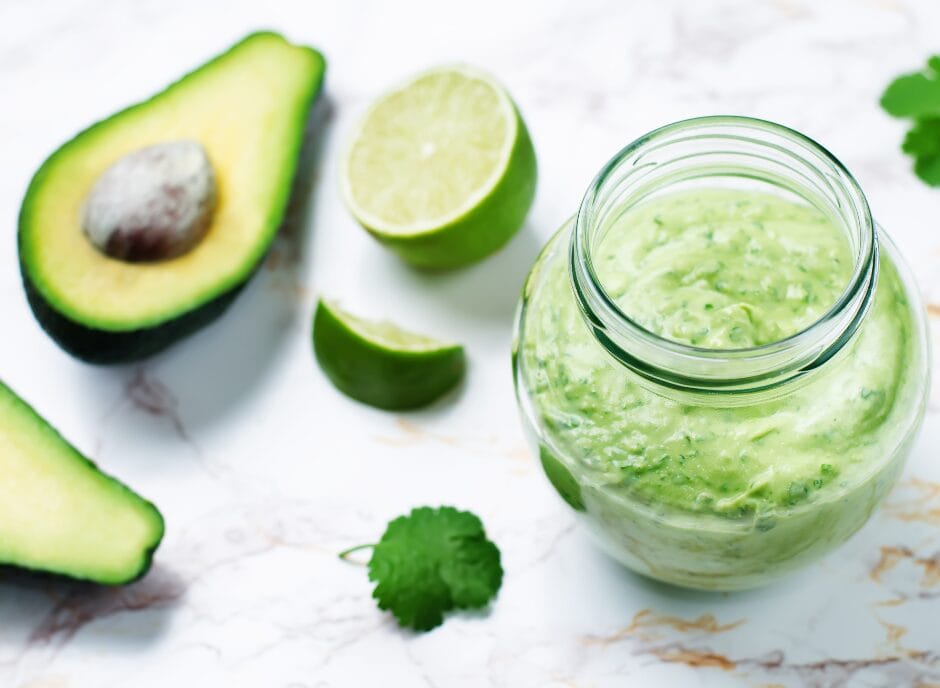A jar of green avocado sauce is surrounded by half an avocado, avocado slices, lime halves, lime wedges, and cilantro leaves on a marble surface.