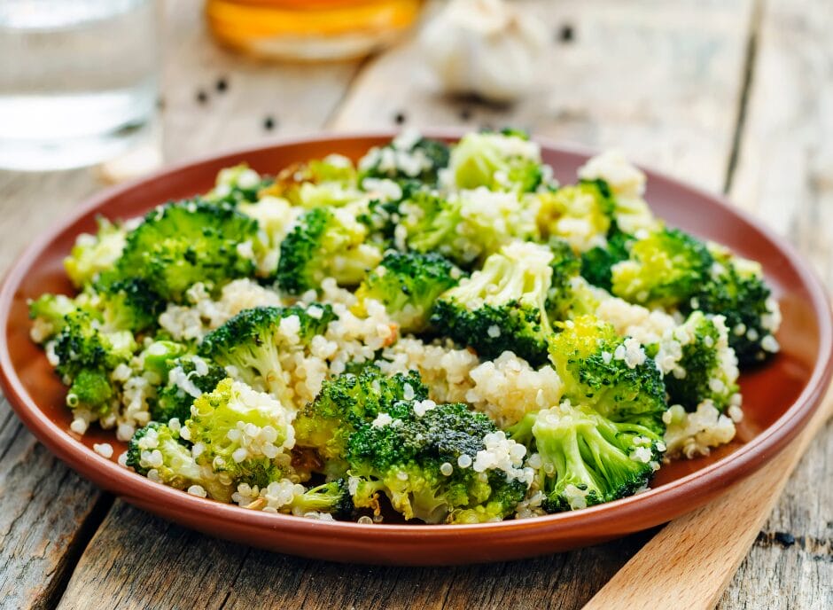 A plate of cooked quinoa and broccoli florets on a wooden surface, with a glass of water and a garlic bulb in the background.