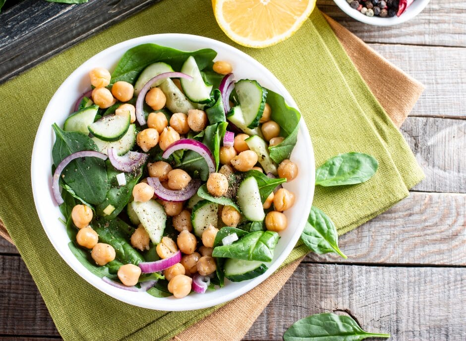 A fresh chickpea salad with spinach, cucumbers, red onions, and a lemon wedge in a white bowl on a wooden table.