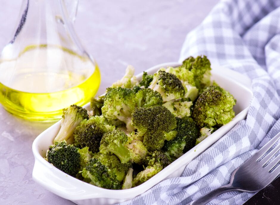 A white dish filled with cooked broccoli next to a clear glass bottle of oil on a checkered cloth.