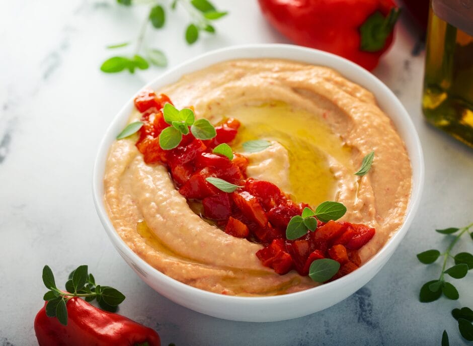 A bowl of creamy red pepper hummus garnished with chopped red peppers, olive oil, and fresh herbs, placed on a white surface.