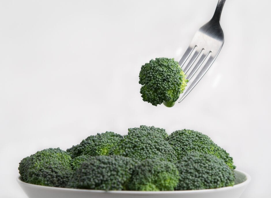 A fork holding a piece of broccoli above a white bowl filled with more broccoli.