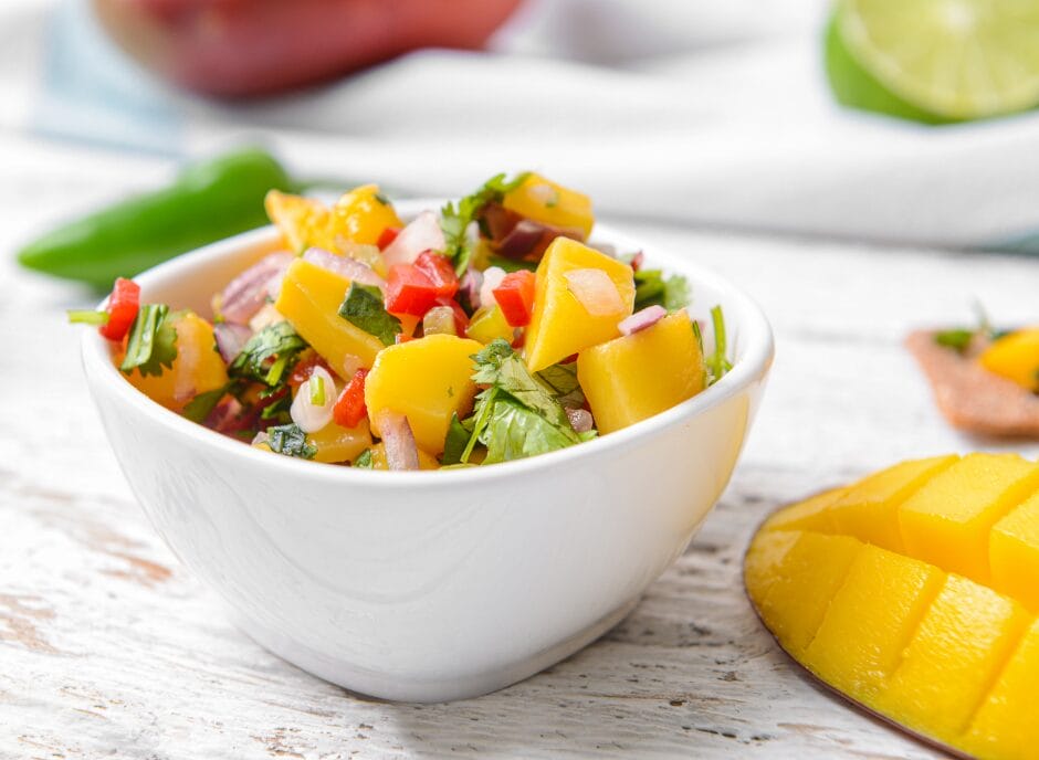 A white bowl filled with mango salsa, including chopped mango, red onion, red bell pepper, cilantro, and other ingredients. A halved lime and sliced mango are in the background.