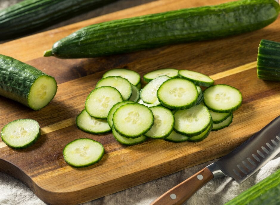 Whole cucumbers and sliced cucumbers on a wooden cutting board, with a knife to the side, on a kitchen counter.