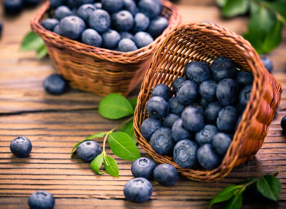 Two wicker baskets filled with fresh blueberries rest on a wooden surface, with a few scattered berries and green leaves nearby.