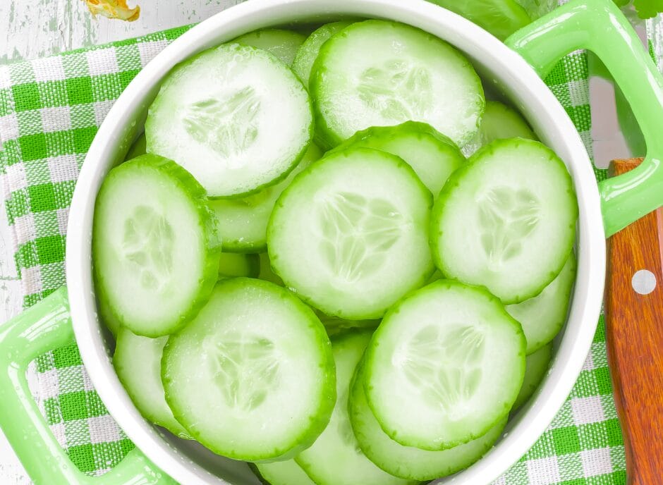 Sliced cucumbers soaking in water in a green pot on a checkered green and white cloth, with a wooden handle visible.