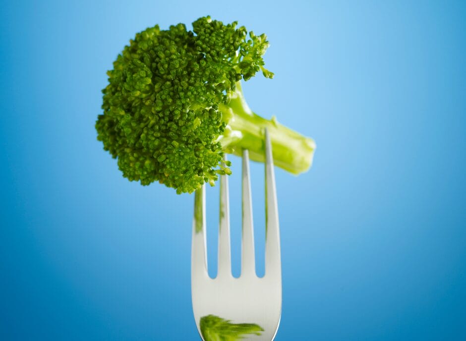 A fork holds a piece of broccoli against a blue background.