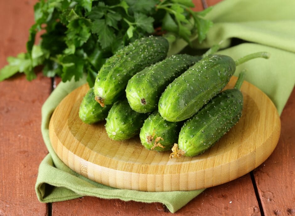 Fresh cucumbers on a wooden cutting board with a green cloth and parsley, on a rustic wooden table.