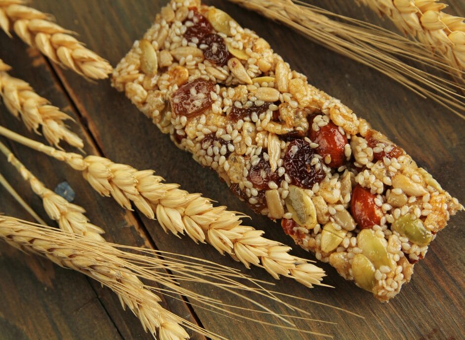 A granola bar with a variety of nuts, seeds, and dried fruits surrounded by stalks of wheat on a wooden surface.