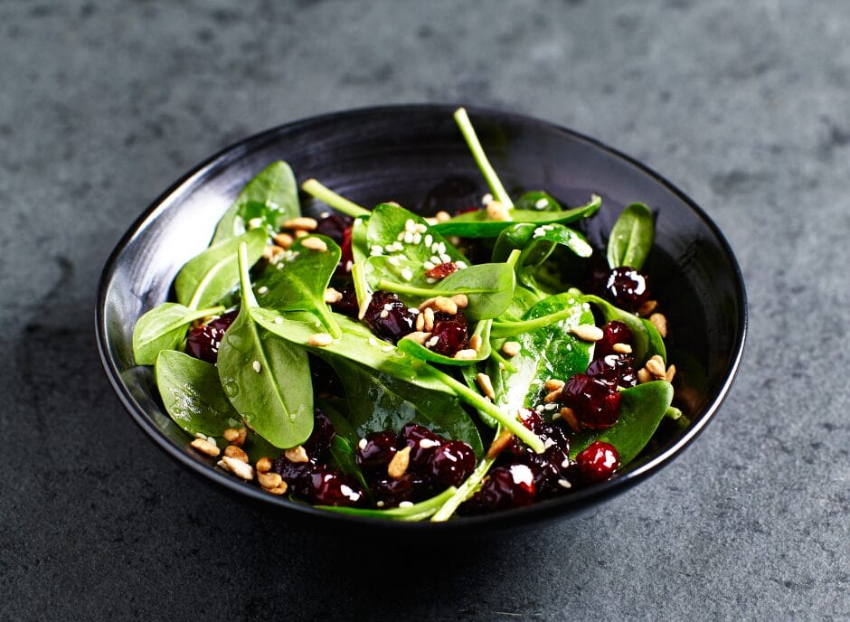 A black bowl of spinach salad with dried cranberries, sunflower seeds, and a light vinaigrette dressing, placed on a dark textured surface.