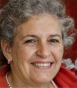 A woman with short curly gray hair and wearing pearl earrings smiles at the camera. She has a red garment draped over her shoulders.