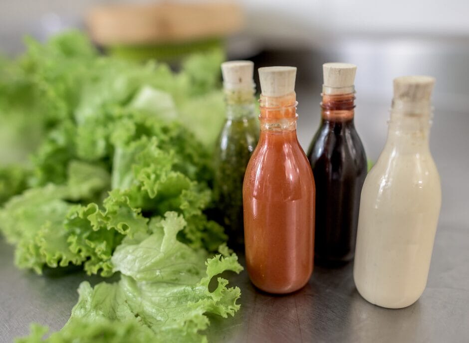 Four bottles of different salad dressings, each with a cork stopper, are placed in front of a bunch of fresh green lettuce leaves on a kitchen countertop.