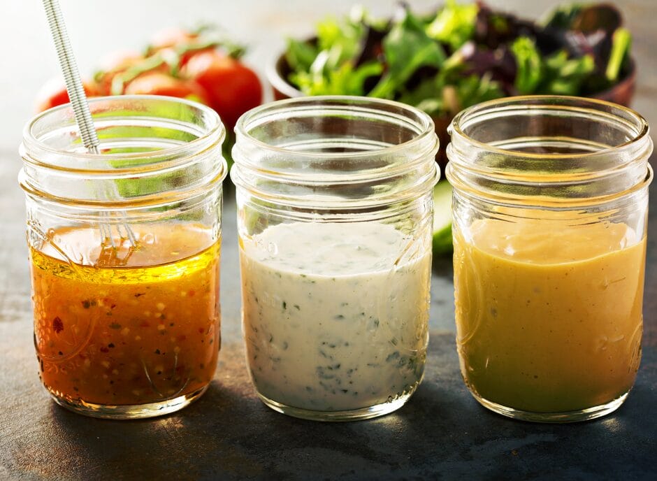 Three glass jars filled with different types of salad dressings are placed on a table with a salad in the background.