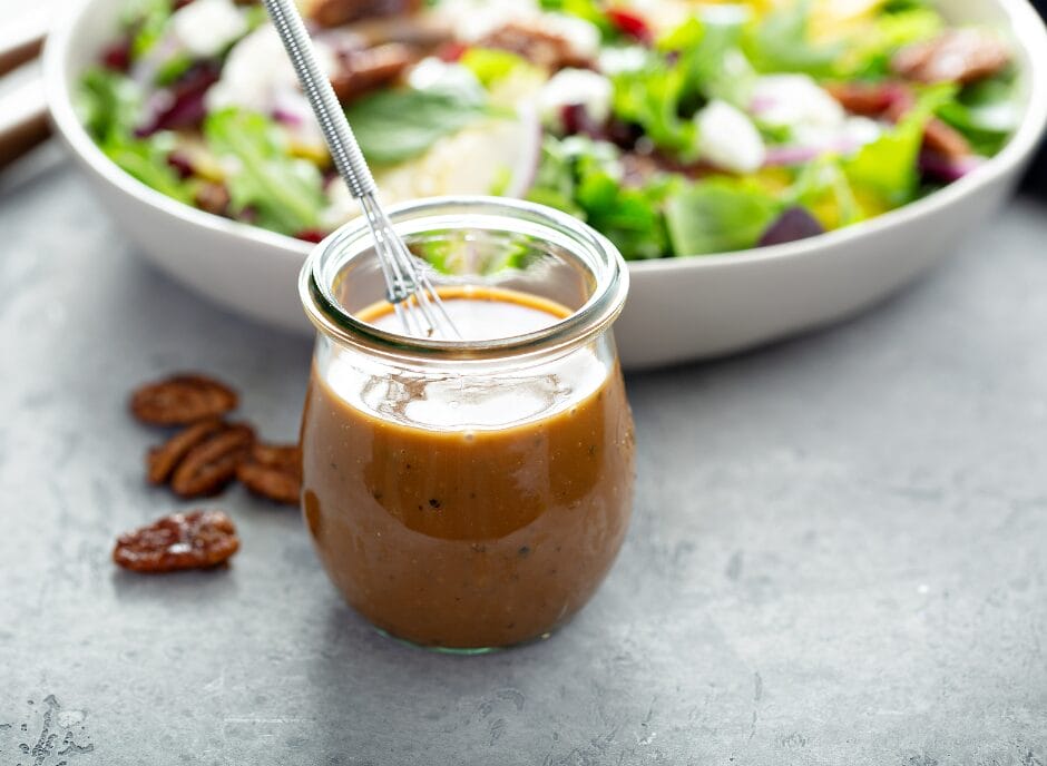 A jar of salad dressing with a stirrer in it is placed in front of a salad bowl on a gray surface.