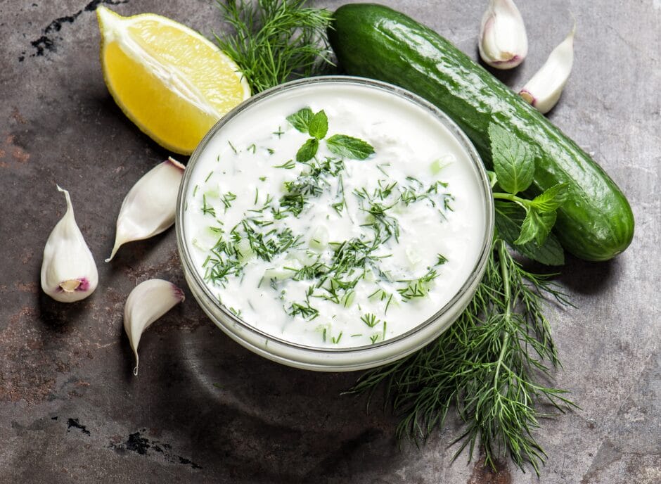 A bowl of tzatziki sauce garnished with dill and mint, surrounded by fresh ingredients: a lemon wedge, cucumber, garlic cloves, and dill sprigs on a rustic surface.