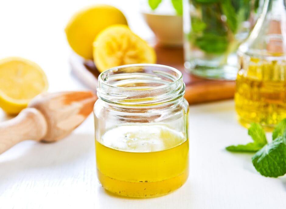 A clear glass jar filled with yellow oil sits on a white surface. In the background, there are lemons, a wooden reamer, and a bottle of oil.