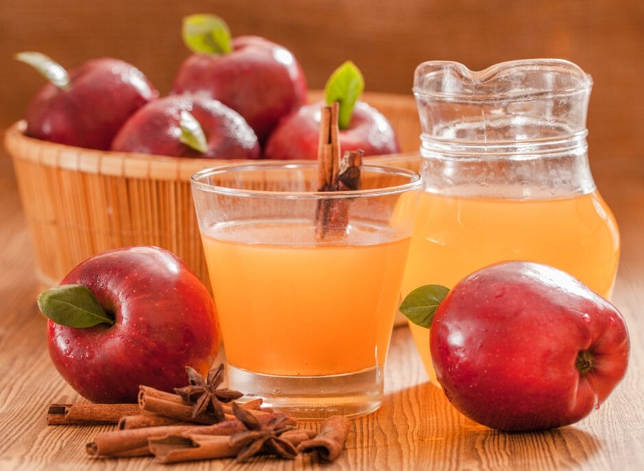 A glass of apple juice with a cinnamon stick, a jug of apple juice, red apples, and a wicker basket filled with apples on a wooden surface.