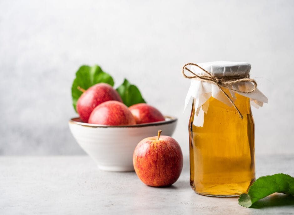 A jar of apple cider vinegar beside a bowl of red apples and a single apple on a light-grey surface.