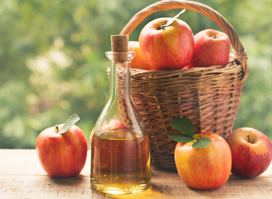 Glass bottle of apple cider vinegar on a wooden surface, next to three apples, with a wicker basket filled with apples in the background.