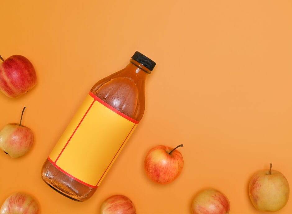 A glass bottle with a blank yellow label lies on an orange background, surrounded by scattered apples.