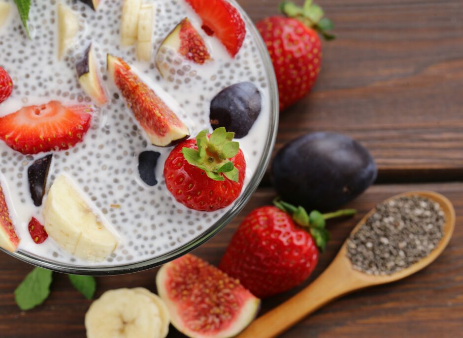 A glass bowl of chia pudding topped with sliced strawberries, bananas, figs, and grapes on a wooden surface. Nearby are fresh strawberries, figs, and a wooden spoon filled with chia seeds.