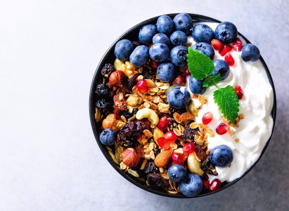 A bowl of yogurt topped with granola, blueberries, pomegranate seeds, and assorted nuts, garnished with fresh mint leaves.