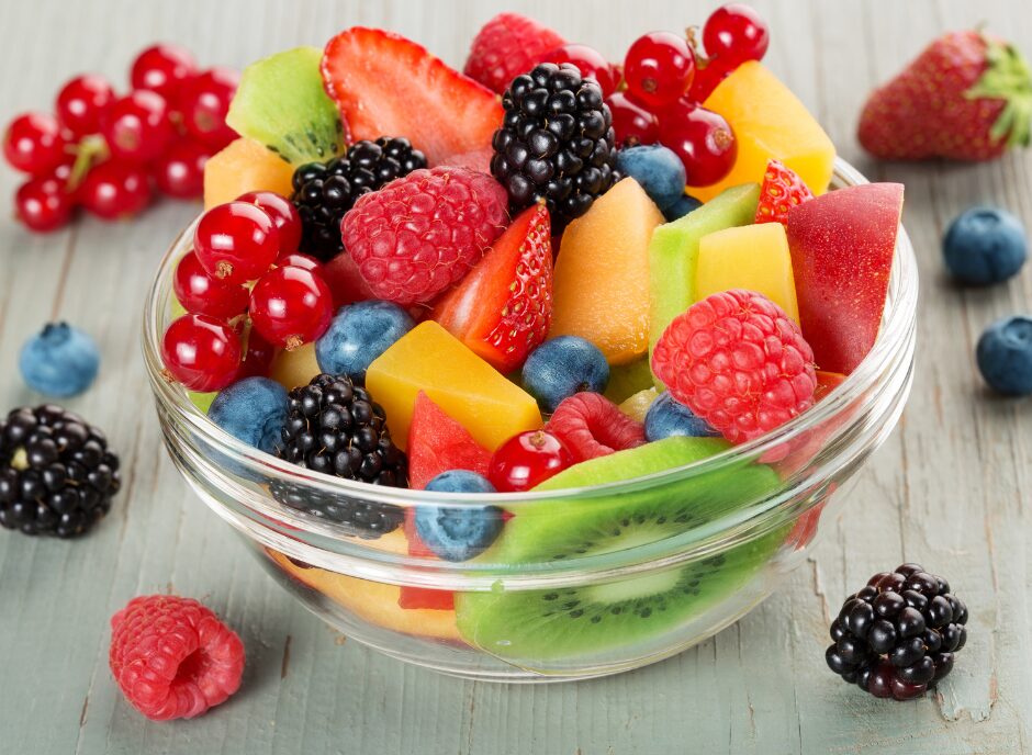 A glass bowl filled with a variety of fresh fruits, including kiwi, raspberries, blackberries, strawberries, blueberries, melon, red currants, and mango, sits on a wooden table.