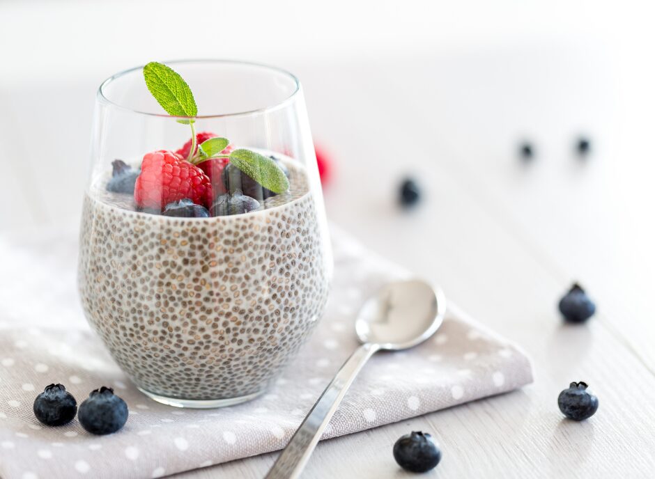 A glass of chia pudding topped with blueberries, raspberries, and a mint leaf, placed on a napkin beside a spoon. Scattered blueberries are also visible on the table.