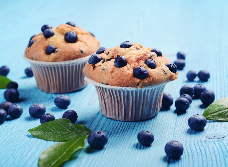 Two blueberry muffins sit on a blue wooden surface, surrounded by fresh blueberries and a few green leaves.