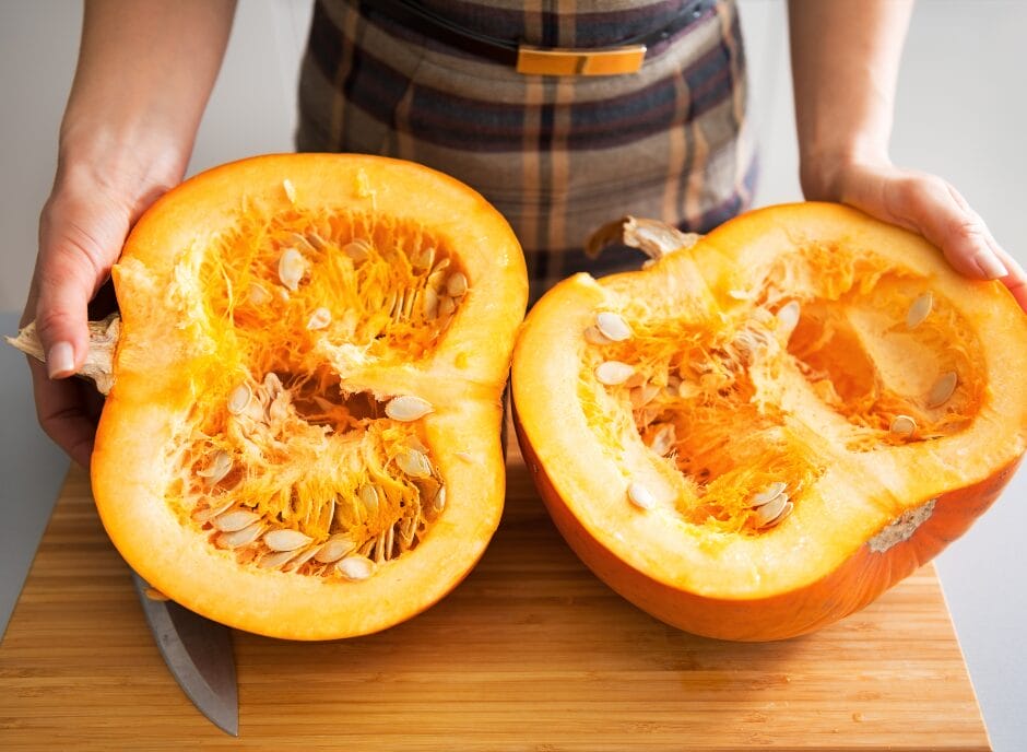 Person holding a halved pumpkin on a wooden board, showing seeds and fibrous interior.