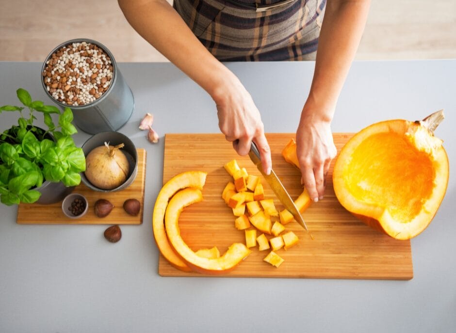 Person slicing pumpkin on a wooden cutting board, with basil, a small pumpkin, nuts, and a plant nearby on a table.