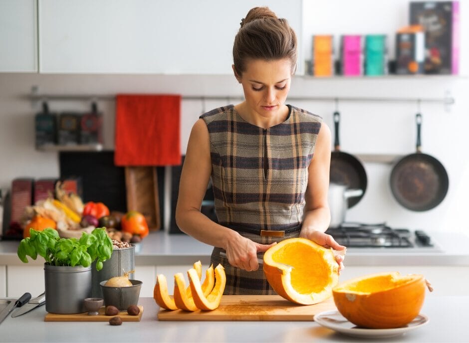 A woman in a kitchen slicing a pumpkin on a cutting board, with kitchen utensils and fresh produce nearby.