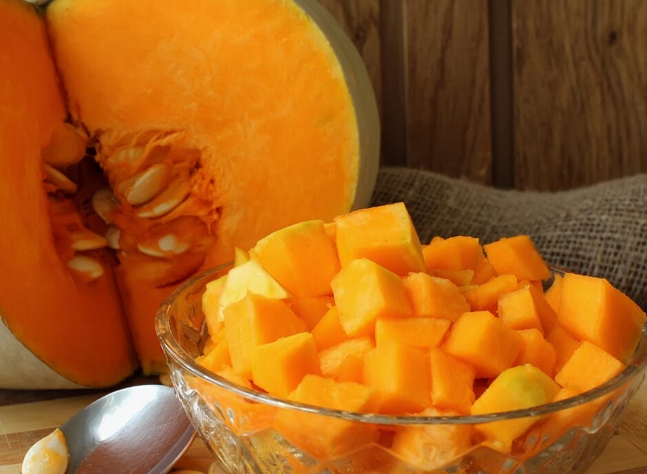 Cubed pumpkin in a glass bowl next to a halved pumpkin with visible seeds.