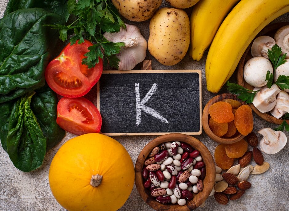 Assorted potassium-rich foods, including bananas, tomatoes, beans, spinach, nuts, mushrooms, potatoes, apricots, and garlic, surround a chalkboard with "K" written on it.