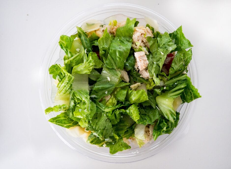 A plastic bowl filled with fresh green salad, featuring romaine lettuce, chunks of chicken, and shredded cheese, viewed from above.