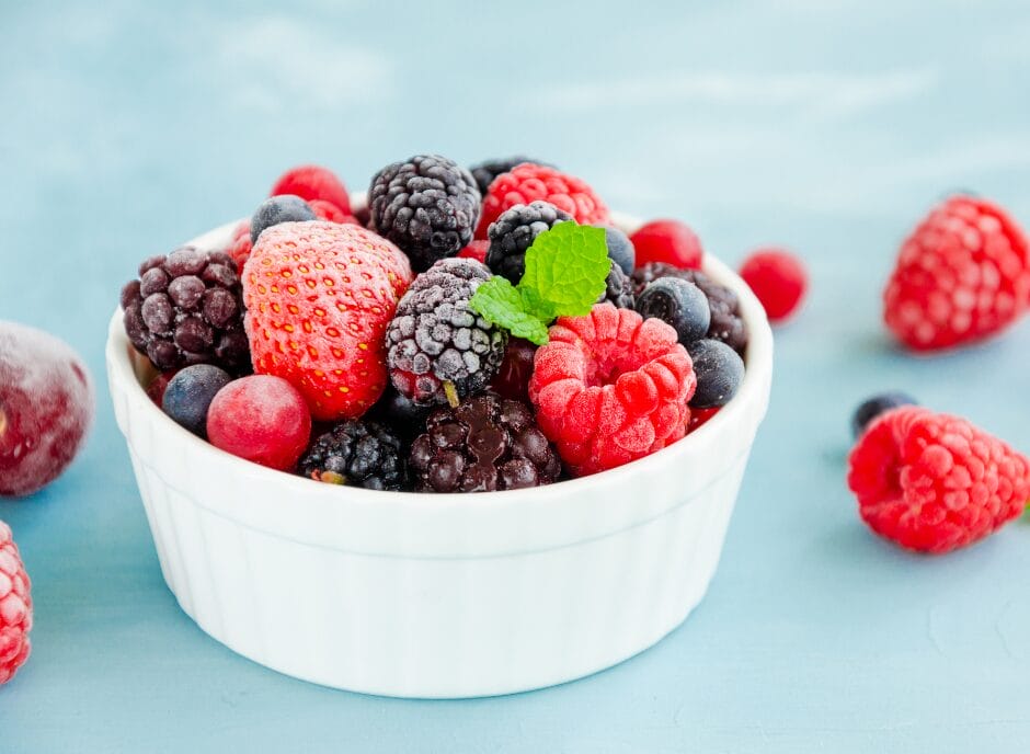 A white bowl filled with assorted frozen berries, including strawberries, raspberries, blackberries, and blueberries, placed on a light blue surface.