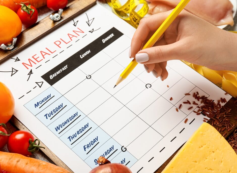 A hand writes on a meal plan surrounded by fresh vegetables, cheese, raw chicken, rice, and a measuring tape.