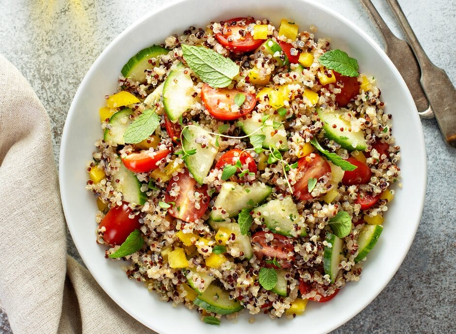 A bowl of quinoa salad with sliced cucumbers, cherry tomatoes, yellow bell peppers, and mint leaves on a textured surface. A napkin and cutlery are beside the bowl.