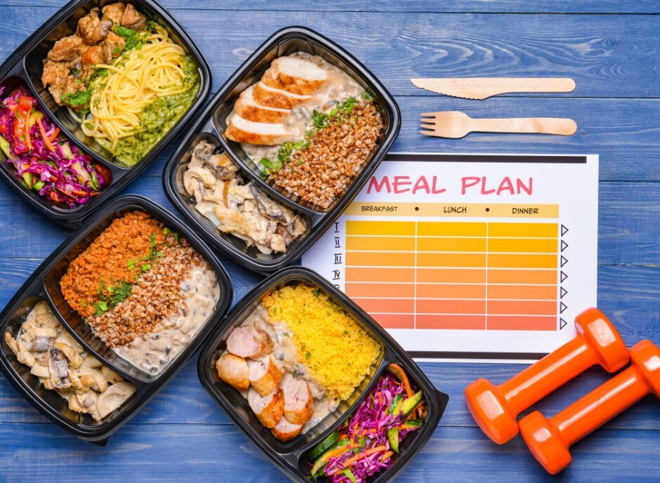 Meal prep containers with various dishes, a meal plan chart, wooden cutlery, and orange dumbbells on a blue surface.