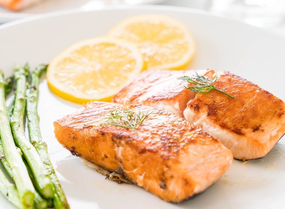 Grilled salmon fillets garnished with herbs, served with asparagus spears and lemon slices on a white plate.